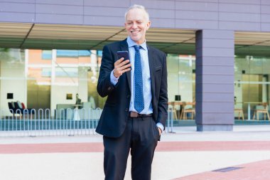 Modern ofis binasının dışındaki akıllı telefonu kullanarak şık takım elbise ve kravat takan kendinden emin kıdemli işadamı. Profesyonel, başarılı ve pozitif tutum kurumsal yaşam tarzını temsil ediyor..