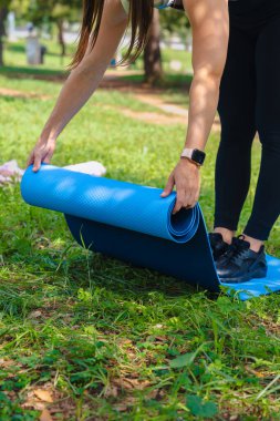 Spor kıyafetleri giyen, yeşil parkta mavi yoga minderi hazırlayan, egzersiz yapmadan önce vücudunu esneten tanınmamış aktif bir kadın. Sağlıklı yaşam tarzı, zindelik, sağlık ve doğa ile denge kavramı.