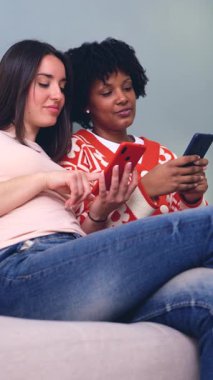 Neşeli Kafkasyalı ve Afro-Amerikalı kadınlar kanepeye oturmuş kırmızı bir akıllı telefona bakarken eğleniyorlar, kahkahaları paylaşıyorlar ve içeride kaliteli zaman geçiriyorlar. Dikey