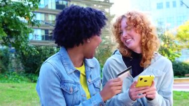  Neşeli Afrikalı Amerikalı ve Kafkasyalı arkadaşlar ellerinde renkli akıllı telefonlar ve bir kredi kartı ile bir şehir parkında duruyorlar. Nakitsiz kolaylığı kutluyorlar, alışveriş başarılarını paylaşıyorlar ve dijital bir yaşam tarzları var.