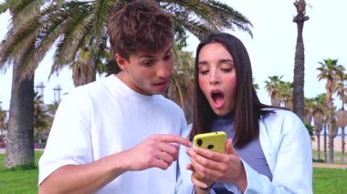 Genç bir adam ve kadın açık havada sarı bir akıllı telefona bakarken gülümsüyor, günlük kıyafetler, arka planda palmiye ağaçları ve mavi gökyüzü, sosyal medyadan birlikte keyif alıyorlar, neşeli ve rahat bir hava. 