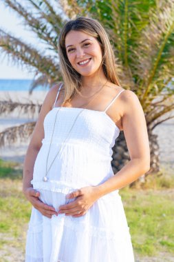 Beyaz yaz elbisesi içinde gülümseyen hamile bir kadının portresi palmiye ağacının yanında durup kameraya bakarken doğal ortam düzeni ve mutluluk sakin bir annelik yaşam tarzını yansıtıyor..