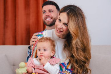 Gülümseyen genç ebeveynler evde bebekle birlikte yerde oturuyorlar, aile sevgisi, mutluluk, ebeveynlik, bağlılık, bakım ve birliktelik kavramı samimi bir aile hayatı, neşeli ve sıcak bir aile anı.