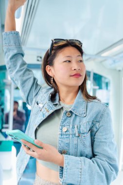 Mutlu Çinli kadın bir elinde tramvay korkuluğuna tutunurken diğer elinde akıllı telefon kullanıyor. Kentsel toplu taşımada gülümsüyor. Yaşam tarzı, teknoloji, seyahat ve işe gidiş gelişler için mükemmel..