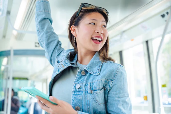 Mutlu Çinli kadın bir elinde tramvay korkuluğuna tutunurken diğer elinde akıllı telefon kullanıyor. Kentsel toplu taşımada gülümsüyor. Yaşam tarzı, teknoloji, seyahat ve işe gidiş gelişler için mükemmel..