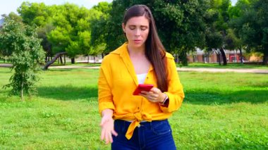 Endişeli kadın, sarı tişörtlü, açık havada akıllı telefona bakan, parkta el üstünde duran, stres konsepti, kötü haber, hayal kırıklığı, hayal kırıklığı, iletişim, teknoloji, duygu.