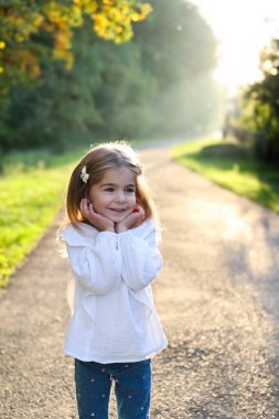 Altın saatinde güneşli bir parkta, elleri yanaklarında gülümseyen mutlu küçük kız. Doğal arka plan ışığı ve yumuşak bokeh. Çocukluk kavramı, masumiyet, özgürlük ve tasasız yaşam tarzı..