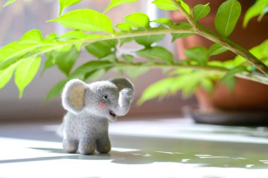 Cute woolly toy elephant under green leaves on a sunny day. Dry felting.