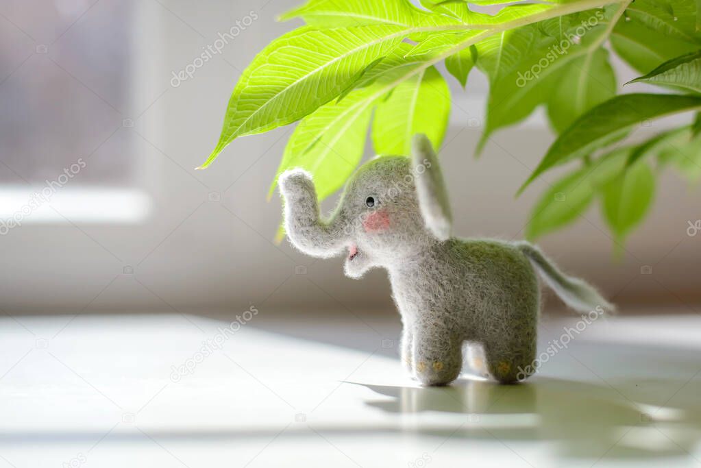 Cute woolly toy elephant under green leaves on a sunny day. Dry felting.