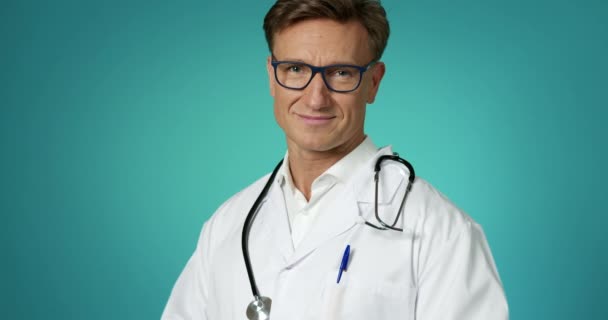 Doctor guapo con una bata blanca de laboratorio con un estetoscopio sonriendo ante la cámara posando en cámara lenta. Profesional Médico en Clinic Looking con Emoción Positiva. Primer plano cara médico trabajador cuidado de la salud Concepto. — Vídeos de Stock