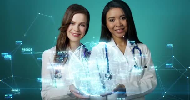 Красивая голограмма, вращающаяся на руках женщин-врачей. Молодые улыбающиеся женщины-медики держат футуристическую цифровую голубую сетевую голограмму, двигающуюся сетку, линии, числа. Концепция современной медицины и технологий — стоковое видео