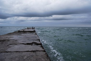 Sonbahar deniz dalgaları beton bir deliğe çarpar. Beton iskele, bulutlu bir gökyüzünün altında fırtınalı deniz dalgalarına karışıp kasvetli bir kıyı manzarası yaratır..