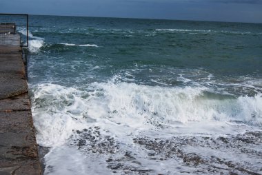 Sonbahar deniz dalgaları beton bir deliğe çarpar. Beton iskele, bulutlu bir gökyüzünün altında fırtınalı deniz dalgalarına karışıp kasvetli bir kıyı manzarası yaratır..