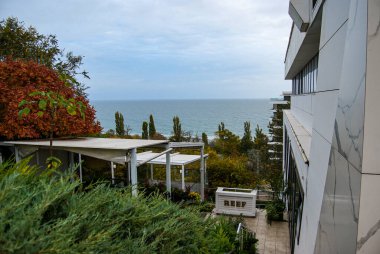 Sonbahar ağaçları ve okyanus ufkuyla kıyı tatil köyü manzarası. Sağında mermer bir ön cephe bulunan gösterişli bir binası olan modern sahil beldesi..