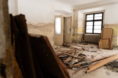 Ahşap döküntüler ve molozlarla dolu terk edilmiş bir odanın içi. Kabuk döken duvarlar, eski bir dökme demir radyatör ve büyük siyah çerçeveli bir pencere.