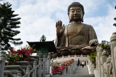 İki ziyaretçi Ngong Ping 'deki Tian Tan Buddha' ya (Büyük Buda) çıkan bougainvillea kaplı merdivene tırmanırken, solda Lantau Adası, Hong Kong 'da bulunan geleneksel bir köşkte bulunan devasa bronz heykel görülüyor.