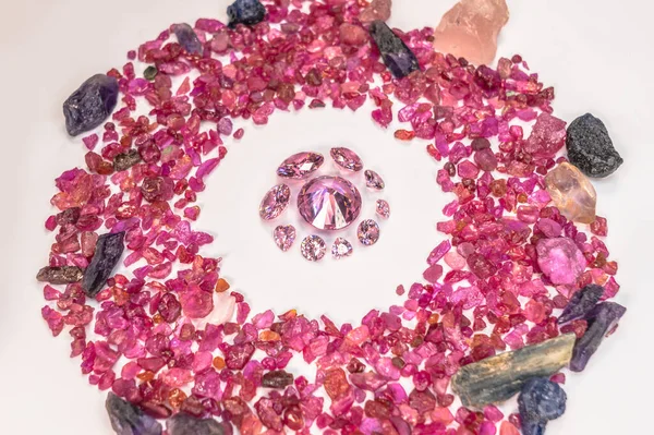 pink sapphires diamonds in different size and many shape are place in the middle on a white ground surrounded by uncut raw red garnet stones