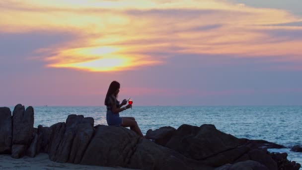 日没の間にビーチでワインとスマートフォンのガラスと美しい若い女性 — ストック動画