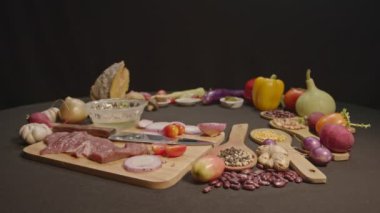 Taze etler, sebzeler ve baharatlar siyah bir masa örtüsüyle kaplanmış bir yemek masasının üzerine serilmiştir..