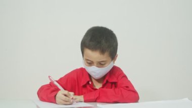 Uzun kollu kırmızı bir gömlek giyen ve maske takan bir çocuk ev ödevini öğretmenine yolluyor. İnternet öğretmenine. Covid-19 salgını yüzünden..