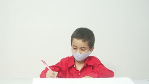 仮面をつけた赤い長袖シャツを着た少年が宿題を座って先生に宿題を送ります インターネット教育Covid 19の流行のため — ストック動画