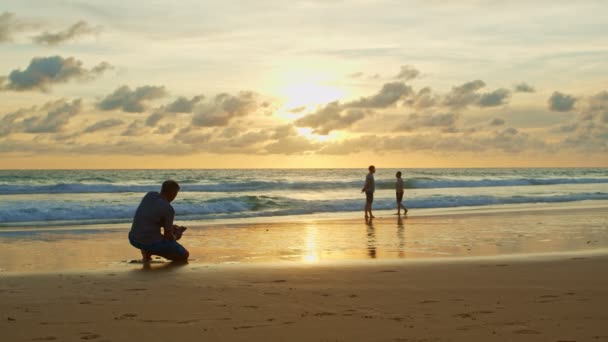 一个男人用手机在海滩上拍照 金色的天空在日落的海面上 视频4K 自然录像高质量的镜头 — 图库视频影像