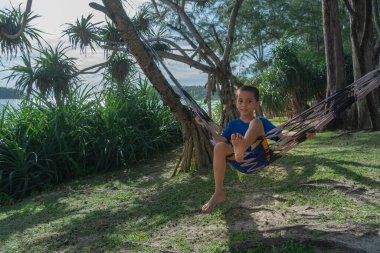 Mavi elbiseli bir çocuk deniz kenarındaki çam ağacının gölgesinde hamakta dinleniyor.