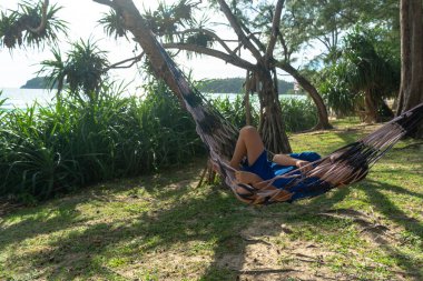 Mavi elbiseli bir çocuk deniz kenarındaki çam ağacının gölgesinde hamakta dinleniyor.