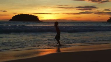 Altın gün batımında beyaz dalgalarda zıplayan bir çocuk. Cennet sahili mavi denizi ve temiz kumsal manzarası. Komik çocuk ve seyahat konsepti.
