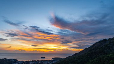 Nefes kesen bir günbatımı, kıyı kasabasının üzerinde turuncu ve mavi renkli canlı renkler çiziyor. Sahneyi çevreleyen yemyeşil tepeler var. Uzak bir tepe yapısı huzur dolu bir manzara ekler..  