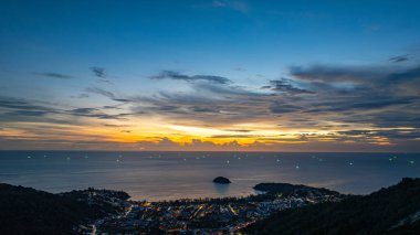 Nefes kesen bir günbatımı, kıyı kasabasının üzerinde turuncu ve mavi renkli canlı renkler çiziyor. Sahneyi çevreleyen yemyeşil tepeler var. Uzak bir tepe yapısı huzur dolu bir manzara ekler..  