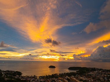 Nefes kesen bir günbatımı, kıyı kasabasının üzerinde turuncu ve mavi renkli canlı renkler çiziyor. Sahneyi çevreleyen yemyeşil tepeler var. Uzak bir tepe yapısı huzur dolu bir manzara ekler..  