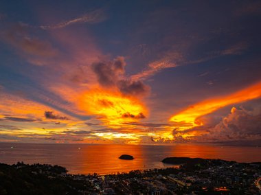 Nefes kesen bir günbatımı, kıyı kasabasının üzerinde turuncu ve mavi renkli canlı renkler çiziyor. Sahneyi çevreleyen yemyeşil tepeler var. Uzak bir tepe yapısı huzur dolu bir manzara ekler..  