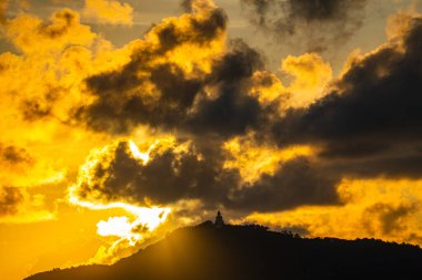 The golden sun sets behind the mountain casting a warm glow around the grand Big Buddha of Phuket. Silhouetted against a radiant sky, the sacred statue radiates peace and spiritual serenity.