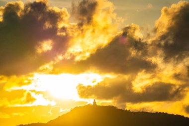 The golden sun sets behind the mountain casting a warm glow around the grand Big Buddha of Phuket. Silhouetted against a radiant sky, the sacred statue radiates peace and spiritual serenity.