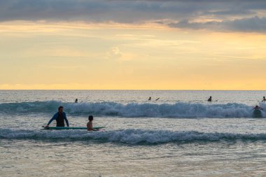 Arkasında yumuşak bir okyanus dalgası yükselirken mor bir sörf tahtasında sörf yapmaktan hoşlanan genç bir çocuk. Sıcak güneş ışığı suyu yansıtıyor, yaz eğlencesinin kaygısız bir anını yakalıyor ve sahilde macera yaşıyor..