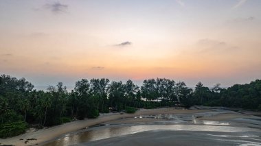 Gün batımında sakin okyanus sularına yansıyan altın rengi ile huzurlu bir sahil adasının çarpıcı hava manzarası. Ağaçların silueti ve kayalık oluşumlar huzur sahnesine derinlik katıyor..
