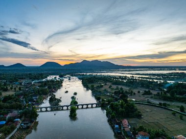 Kırsal tarım arazileri ve köylerden akan geniş bir nehrin manzaralı bir manzarası, suyu geçen bir köprü ve yumuşak bir günbatımı gökyüzüne karşı silueti olan dağlar. Görüntü sükuneti, doğal güzelliği ve peyzaj arasındaki uyumu aktarır.