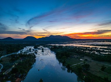 Kırsal tarım arazileri ve köylerden akan geniş bir nehrin manzaralı bir manzarası, suyu geçen bir köprü ve yumuşak bir günbatımı gökyüzüne karşı silueti olan dağlar. Görüntü sükuneti, doğal güzelliği ve peyzaj arasındaki uyumu aktarır.