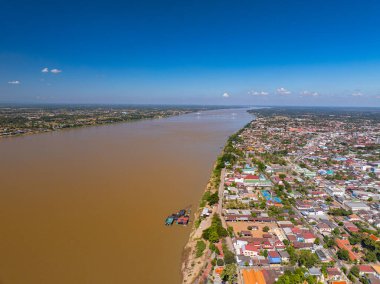 Güneşin sıcacık parıltısıyla aydınlanan nehir kenarındaki bir şehrin çarpıcı hava manzarası. Kalın bulutların dokulu gökyüzü altında altın ışık yayılıyor. Nehir kıyısı, binalar ve canlı ufuk dinamik ve atmosferik bir manzara yaratır..