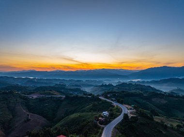 Şafak sökerken, güneş ışığı ormanlık tepelerin üzerine düşer, gökyüzünü turuncu ve mavinin hassas eğimleriyle boyardı. Zaman atlaması, sakin bir dağ manzarasında sessizce uyanan doğayı yakalar.