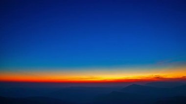 Şafak sökerken, güneş ışığı ormanlık tepelerin üzerine düşer, gökyüzünü turuncu ve mavinin hassas eğimleriyle boyardı. Zaman atlaması, sakin bir dağ manzarasında sessizce uyanan doğayı yakalar..