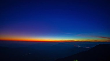 Şafak sökerken, güneş ışığı ormanlık tepelerin üzerine düşer, gökyüzünü turuncu ve mavinin hassas eğimleriyle boyardı. Zaman atlaması, sakin bir dağ manzarasında sessizce uyanan doğayı yakalar..
