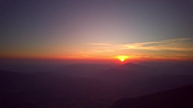 Sisli bir dağ vadisini aydınlatan altın bir güneşin sinematik hava görüntüsü. Yumuşak sabah ışığı sislerden oluşan bir denize yayılır, ufuktaki uzak sıradağları ortaya çıkarır, doğa yolculuğu