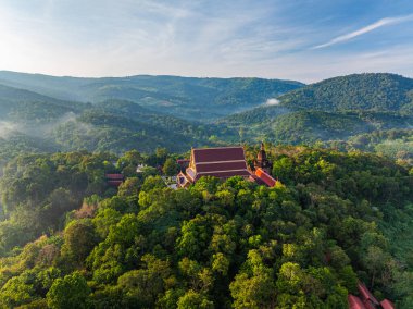 Bereketli yeşil orman dağları arasında yuva yapmış dingin bir tapınağın havadan görünüşü, berrak mavi bir gökyüzü altında ruhani mimari ve çevresindeki doğal manzara arasındaki uyumu yakalıyor..
