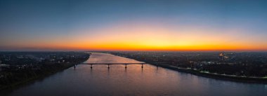 Gün batımında geniş bir nehri kaplayan bir şehir köprüsünün havadan görünüşü. Yumuşak altın ışık ve renkli gökyüzü suya yansıyor. Görüntü, sükunet, seyahat ve şehir manzarasını gece gündüz aktarır.