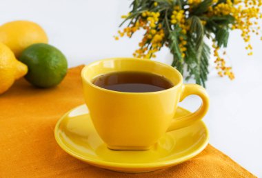 Sarı fincanda siyah çay ve parlak bir mimoza dalı.