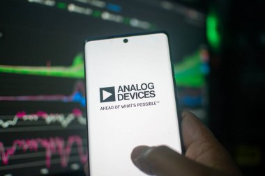 Dhaka, Bangladeş 20 Şubat 2026: Analog Aygıtları logosunu borsa grafiklerinin önünde gösteren akıllı telefonu tutan bir el