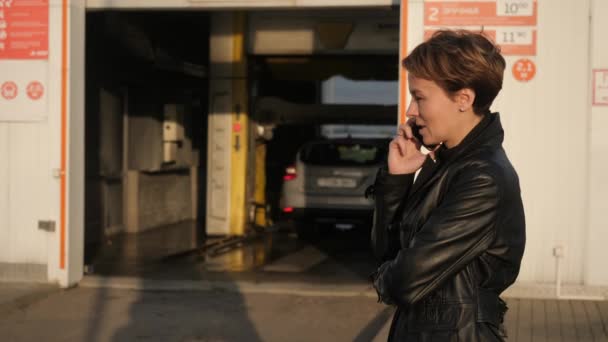 Ελκυστική κοπέλα που μιλάει στο τηλέφωνο στο παρασκήνιο ενός πλυντηρίου αυτοκινήτων. — Αρχείο Βίντεο