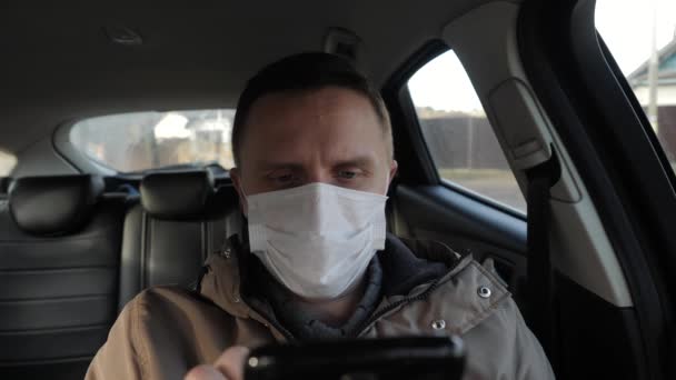 Mężczyzna w masce medycznej siedzi w samochodzie i używa smartfona.. — Wideo stockowe